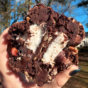 Puede incluir: Primer plano de una galleta brownie de chocolate con relleno de malvavisco y nueces. La galleta está partida por la mitad, revelando el malvavisco pegajoso en el interior.