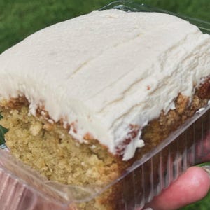 Può includere: Una fetta di torta con un spesso strato di glassa bianca. La torta ha una base marrone chiaro ed è in un contenitore di plastica trasparente. La torta sembra umida e deliziosa.