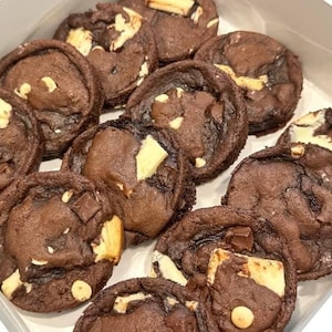 May include: A box filled with freshly baked chocolate cookies. The cookies are dark brown with chunks of white chocolate and dark chocolate pieces. The cookies are arranged in a white box.