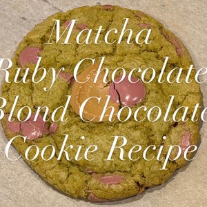 May include: A close-up of a matcha cookie with ruby and blond chocolate chips. The cookie has a light green hue and is speckled with pink and tan chocolate pieces. Text on the cookie reads "Matcha Ruby Chocolate Blond Chocolate Cookie Recipe."