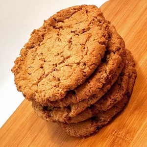 以下が含まれることがあります： 木製のまな板の上に、薄い茶色のクッキーが4枚重ねられています。クッキーは丸く、少しざらざらした質感です。