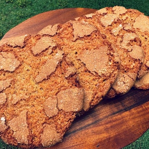 May include: Four round loaves of brown bread with a slightly crispy crust and a rough, textured surface. The bread is arranged in a stack on a wooden chopping board.