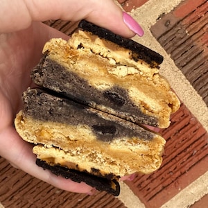May include: A close-up of a cookie sandwich with a chocolate cookie base and a peanut butter cookie top. The cookie is cut in half to show the layers.
