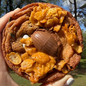 May include: A large, round cookie with a textured, golden-brown exterior. The cookie is topped with crunchy corn flakes, a chocolate egg, and a caramel-like glaze. A hand holds the cookie against a blurred outdoor background.