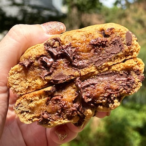 Puede incluir: Primer plano de una galleta de chocolate con chips con relleno de caramelo y chocolate. La galleta está partida por la mitad, revelando el relleno pegajoso en el interior.