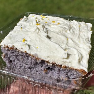 May include: A square slice of purple cake topped with a thick layer of white frosting and yellow sprinkles. The cake is in a clear plastic container.