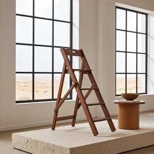 Könnte beinhalten: Ein klappbarer Trittschemel aus dunkelbraunem Holz mit drei Stufen. Der Hocker steht auf einer strukturierten, hellfarbigen Oberfläche. Der Hintergrund zeigt große Fenster mit schwarzen Rahmen und einen kleinen Beistelltisch aus Holz. Die Höhe beträgt ca. 91 cm.