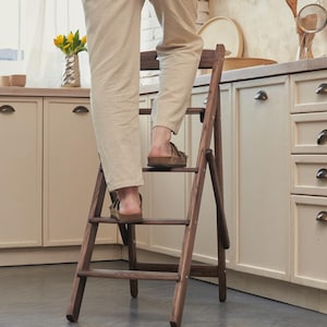 May include: A wooden folding step stool with three steps. The stool is brown and appears to be in a kitchen setting. The person is standing on the stool. The kitchen has cream-colored cabinets and a window.