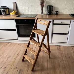 May include: A wooden folding step stool with three steps and a seat. The stool is made of light-colored wood with a natural grain pattern. It is positioned in a kitchen setting, suggesting its use for reaching high shelves or cabinets.