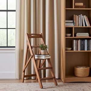 Peut inclure: Un escabeau pliant en bois à trois marches, doté d'une finition brun moyen. Une petite plante verte en pot est posée sur la marche supérieure, sur deux livres blancs empilés. L'escabeau présente une poignée découpée et des supports latéraux.