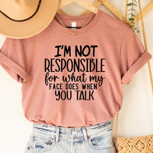 May include: Heather mauve t-shirt with the text "I'M NOT RESPONSIBLE for what my FACE DOES WHEN YOU TALK". The shirt is paired with denim shorts, a tan hat, and a woven crossbody bag. The shirt is hanging on a wooden hanger.