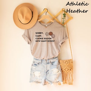 Pu&ograve; includere: Una maglietta grigio chiaro con la scritta "SORRY. CANT. COOKIE SEASON. HOW MANY BOXES?" e illustrazioni di biscotti. La maglietta &egrave; abbinata a pantaloncini di jeans, una borsa intrecciata e un cappello beige. Le parole "Athletic Heather" sono nell'angolo in alto a destra.