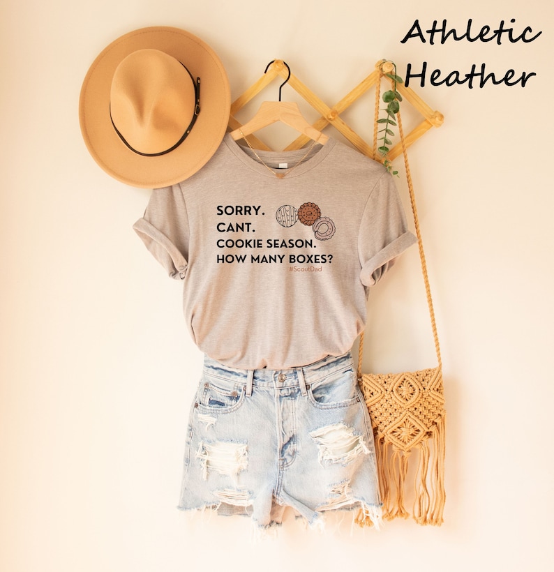 Pu&ograve; includere: Una maglietta grigio chiaro con la scritta "SORRY. CANT. COOKIE SEASON. HOW MANY BOXES?" e illustrazioni di biscotti. La maglietta &egrave; abbinata a pantaloncini di jeans, una borsa intrecciata e un cappello beige. Le parole "Athletic Heather" sono nell'angolo in alto a destra.