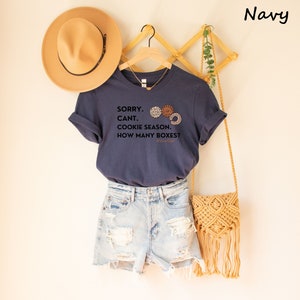 Pu&ograve; includere: T-shirt blu navy con la scritta "SORRY. CANT. COOKIE SEASON. HOW MANY BOXES?" e grafica di biscotti. La maglietta &egrave; abbinata a pantaloncini di jeans, un cappello beige e una borsa intrecciata. La maglietta &egrave; appesa a una gruccia di legno.