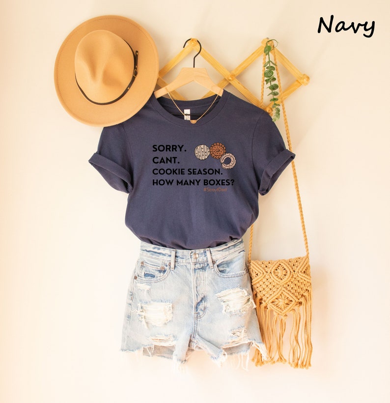 Pu&ograve; includere: T-shirt blu navy con la scritta "SORRY. CANT. COOKIE SEASON. HOW MANY BOXES?" e grafica di biscotti. La maglietta &egrave; abbinata a pantaloncini di jeans, un cappello beige e una borsa intrecciata. La maglietta &egrave; appesa a una gruccia di legno.