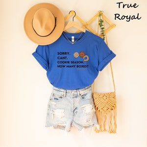 Pu&ograve; includere: Una maglietta blu reale con la scritta "SORRY. CANT. COOKIE SEASON. HOW MANY BOXES?" e grafica di biscotti. La maglietta &egrave; abbinata a pantaloncini di jeans e un cappello color cachi. Una borsa in macram&egrave; &egrave; appesa nelle vicinanze.