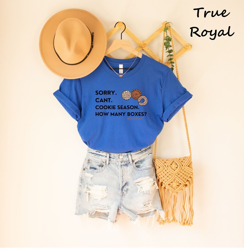 Pu&ograve; includere: Una maglietta blu reale con la scritta "SORRY. CANT. COOKIE SEASON. HOW MANY BOXES?" e grafica di biscotti. La maglietta &egrave; abbinata a pantaloncini di jeans e un cappello color cachi. Una borsa in macram&egrave; &egrave; appesa nelle vicinanze.