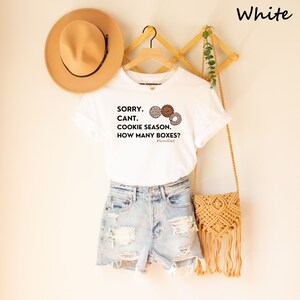 Pu&ograve; includere: T-shirt bianco con la scritta "SORRY. CANT. COOKIE SEASON. HOW MANY BOXES?" e illustrazioni di biscotti. La maglietta &egrave; abbinata a pantaloncini di jeans, un cappello beige e una borsa a tracolla intrecciata.