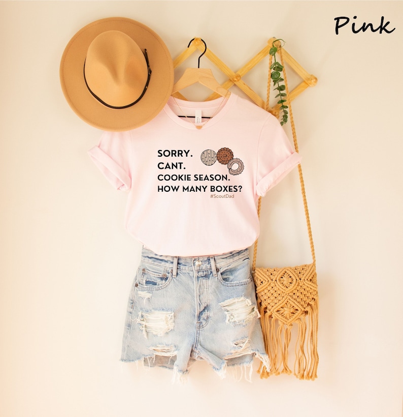 Pu&ograve; includere: Una maglietta rosa chiaro con scollo a V con la scritta "SORRY. CANT. COOKIE SEASON. HOW MANY BOXES?" e illustrazioni di biscotti. La maglietta &egrave; abbinata a pantaloncini di jeans, un cappello beige e una borsa intrecciata.