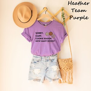 Pu&ograve; includere: Una maglietta viola melange con maniche arrotolate, con la scritta "SORRY. CANT. COOKIE SEASON. HOW MANY BOXES?" e grafica di biscotti. La maglietta &egrave; abbinata a pantaloncini di jeans, un cappello marrone e una borsa intrecciata.
