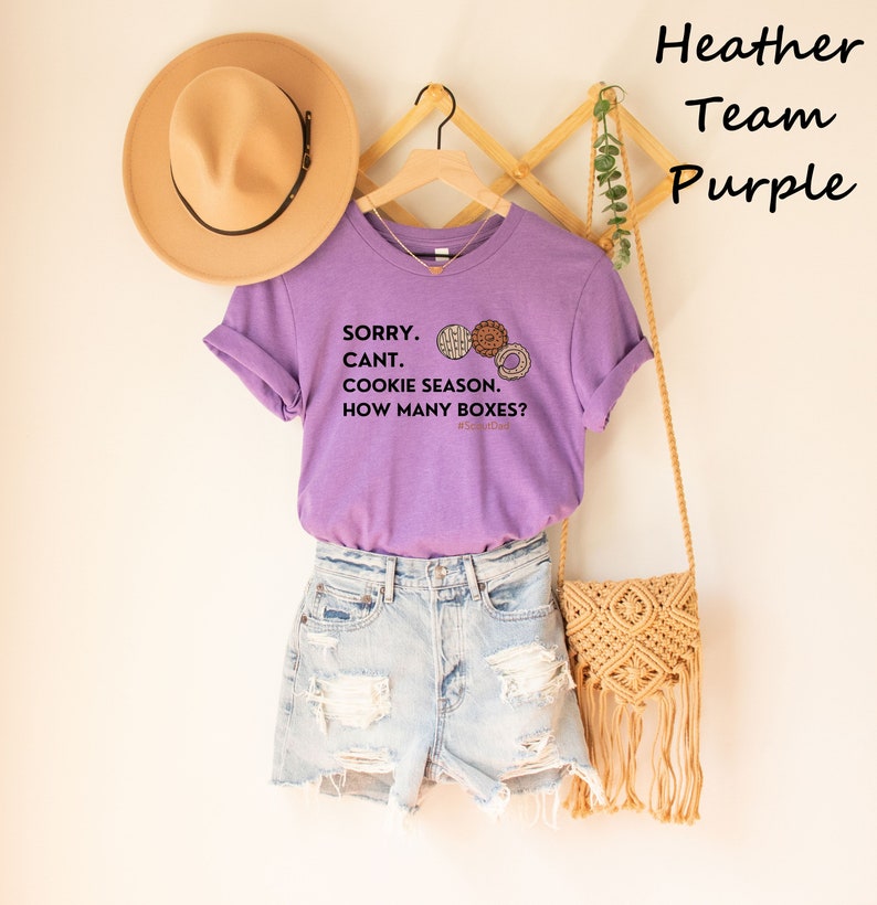 Pu&ograve; includere: Una maglietta viola melange con maniche arrotolate, con la scritta "SORRY. CANT. COOKIE SEASON. HOW MANY BOXES?" e grafica di biscotti. La maglietta &egrave; abbinata a pantaloncini di jeans, un cappello marrone e una borsa intrecciata.