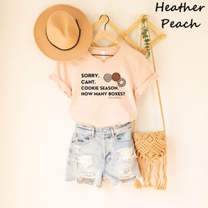 Pu&ograve; includere: Una maglietta color pesca con la scritta "SORRY. CANT. COOKIE SEASON. HOW MANY BOXES?" e illustrazioni di biscotti. La maglietta &egrave; abbinata a pantaloncini di jeans, un cappello beige e una borsa intrecciata.