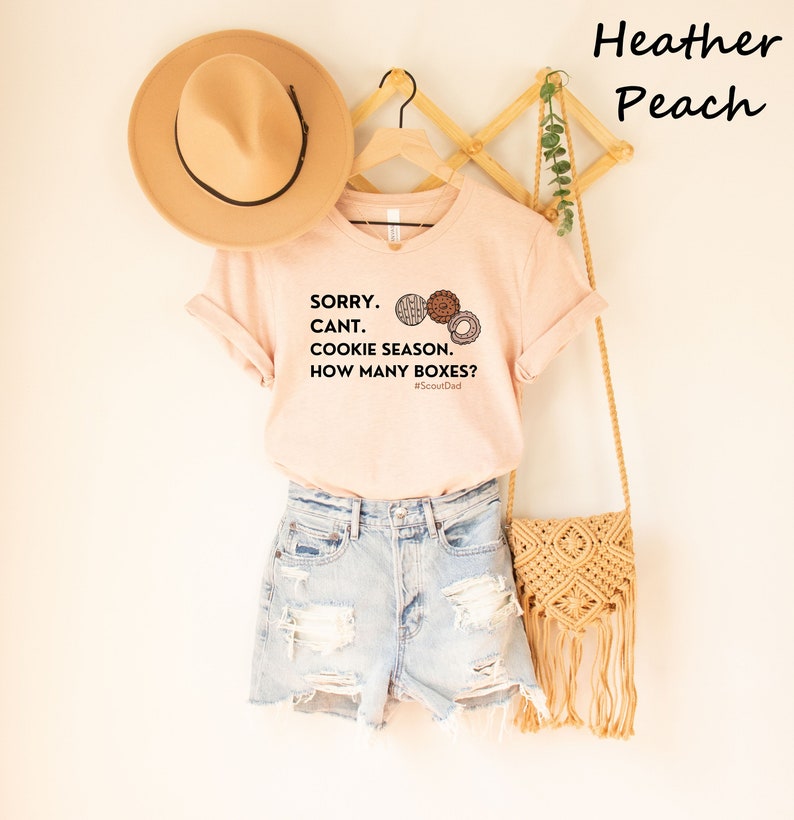 Pu&ograve; includere: Una maglietta color pesca con la scritta "SORRY. CANT. COOKIE SEASON. HOW MANY BOXES?" e illustrazioni di biscotti. La maglietta &egrave; abbinata a pantaloncini di jeans, un cappello beige e una borsa intrecciata.