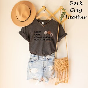 Pu&ograve; includere: T-shirt grigio scuro melange con la scritta "SORRY. CANT. COOKIE SEASON. HOW MANY BOXES?" e tre grafiche di biscotti. La maglietta &egrave; abbinata a pantaloncini di jeans, un cappello beige e una borsa in macram&egrave;.