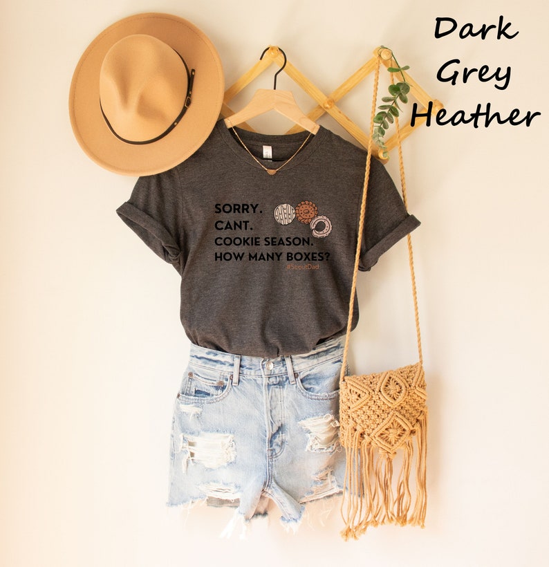 Pu&ograve; includere: T-shirt grigio scuro melange con la scritta "SORRY. CANT. COOKIE SEASON. HOW MANY BOXES?" e tre grafiche di biscotti. La maglietta &egrave; abbinata a pantaloncini di jeans, un cappello beige e una borsa in macram&egrave;.