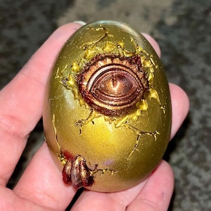 May include: A gold egg with a cracked surface and a red eye peering out. The egg has a metallic sheen and is decorated with gold and red accents.