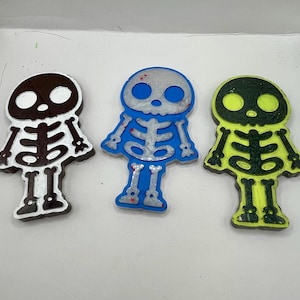 May include: Three resin skeleton ornaments in different colors. The skeletons are black, white, and green with glitter accents. They are all wearing a simple skeleton costume.