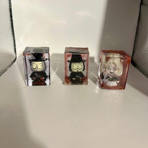 May include: Three small resin figurines encased in clear cubes. The figurines are of a person wearing a black hat and mask, another person wearing a black hat and mask, and a person with blonde hair wearing a white dress.
