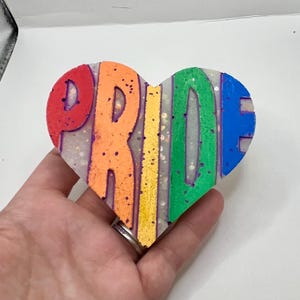 May include: A heart-shaped resin ornament with the word "PRIDE" spelled out in rainbow colors. The letters are outlined in purple and the background is white with glitter.