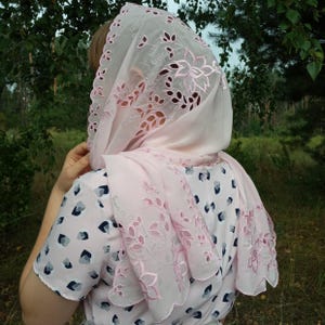 以下が含まれることがあります： 花と葉の切り抜きとスカラップの縁取りが施された、薄いピンク色のヘッドスカーフ。スカーフは頭と肩にかけられています。その人は、青と黒の模様の入った薄いピンク色のドレスを着ています。