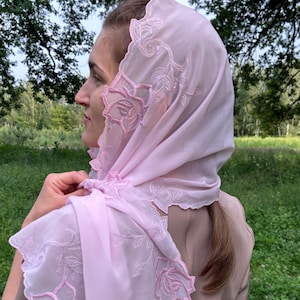 以下が含まれることがあります： 刺繍された花と葉のデザインが施された、薄いピンク色のヘッドスカーフ。スカーフは頭と肩にかけられ、刺繍のディテールは少し濃いピンク色です。スカーフの端はスカラップトリムになっています。