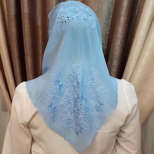 以下が含まれることがあります： 花柄刺繍が施された水色のヘッドカバー。正方形のスカーフは、縁と中央に刺繍された花と葉が特徴です。生地は透けており、縁はスカラップになっています。