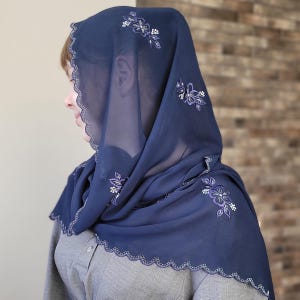 以下が含まれることがあります： 刺繍された花柄とスカラップエッジが特徴の、ネイビーブルーのヘッドカバー。 薄手の生地が頭と肩を覆い、繊細な白と紫の花の刺繍が施されています。 ヘッドカバーはグレーの衣服の上から着用されています。