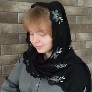 May include: Black head covering with white floral embroidery. The head covering has a scalloped edge and is draped over the head and shoulders. The background is a brick wall.