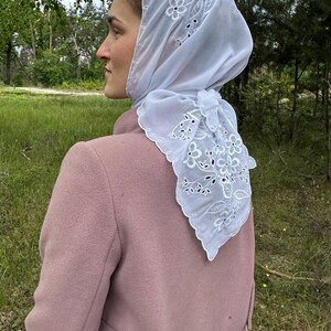 May include: A white, sheer headscarf with floral embroidery and a scalloped edge. The scarf is tied at the back of the neck and worn over a pink coat. The background features green grass and trees.