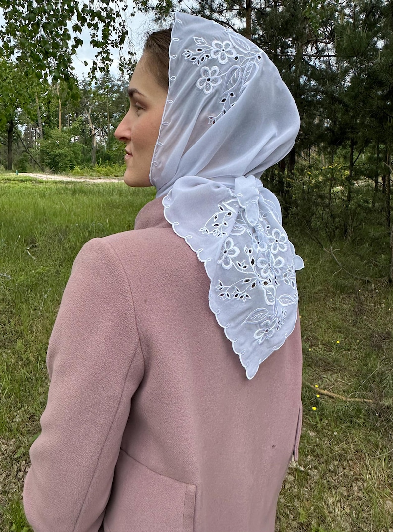 May include: A white, sheer headscarf with floral embroidery and a scalloped edge. The scarf is tied at the back of the neck and worn over a pink coat. The background features green grass and trees.