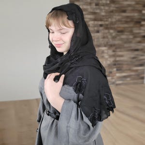 以下が含まれることがあります： 黒いヘッドカバー、花の刺繍入り。 シアー生地が頭と肩にドレープします。 ヘッドカバーはグレーのドレスの上に着用されています。