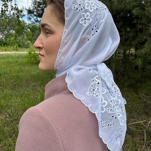 May include: A white, sheer headscarf with floral embroidery and a scalloped edge. The scarf is draped over a person's head and shoulders, with the design visible. The person is wearing a pink coat, and the background is a natural outdoor setting.