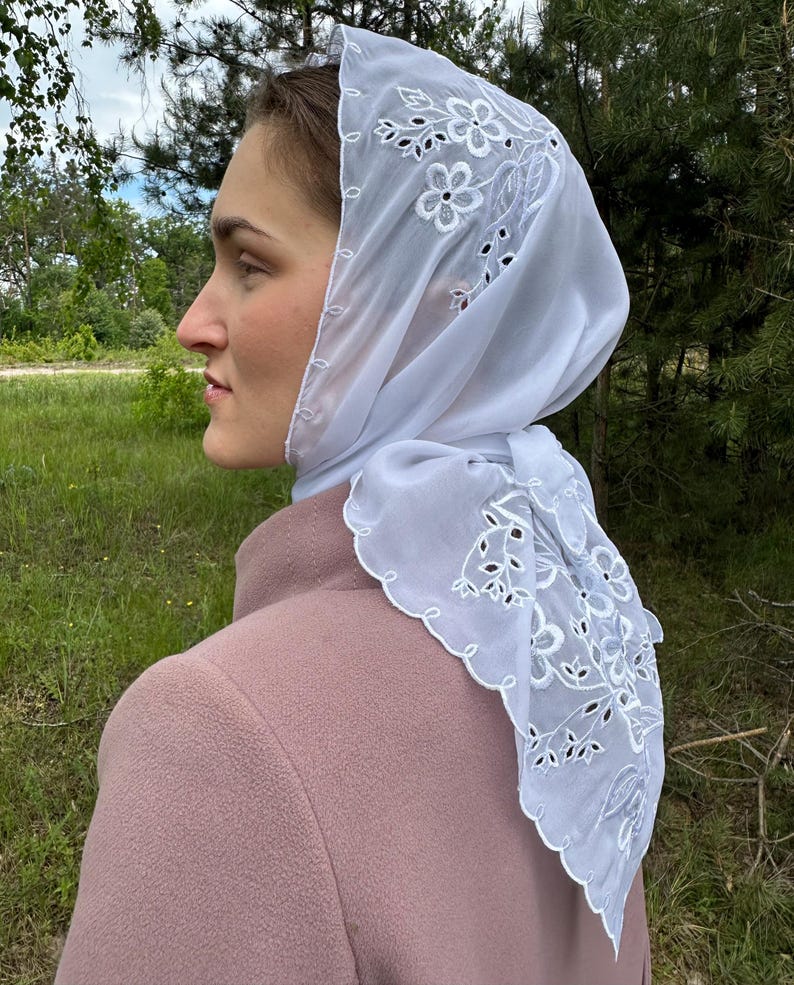 May include: A white, sheer headscarf with floral embroidery and a scalloped edge. The scarf is draped over a person's head and shoulders, with the design visible. The person is wearing a pink coat, and the background is a natural outdoor setting.