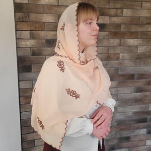 以下が含まれることがあります： ピーチ色のヘッドカバーで、スカラップエッジと刺繍された花の模様があります。 シアー生地が肩にドレープします。 ヘッドカバーは白いブラウスの上に着用されています。