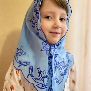 以下が含まれることがあります： 花柄の刺繍が施された薄い青色のスカーフをつけた若い女の子。スカーフはフリルのある縁取りで、透き通った生地で作られています。