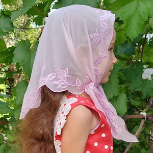 以下が含まれることがあります： 淡いピンク色のシアーヘッドスカーフ。お揃いのピンク色の刺繍の花柄が施されています。スカーフはスカラップエッジで、頭にかけられています。背景には緑の葉と赤い水玉模様のドレスがあります。