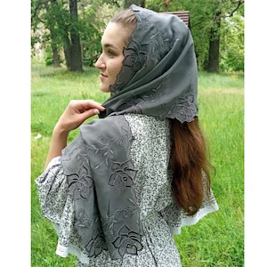 以下が含まれることがあります： 花柄の刺繍が施されたグレーのシアーなスカーフ。スカーフは人の頭と肩にかけられています。