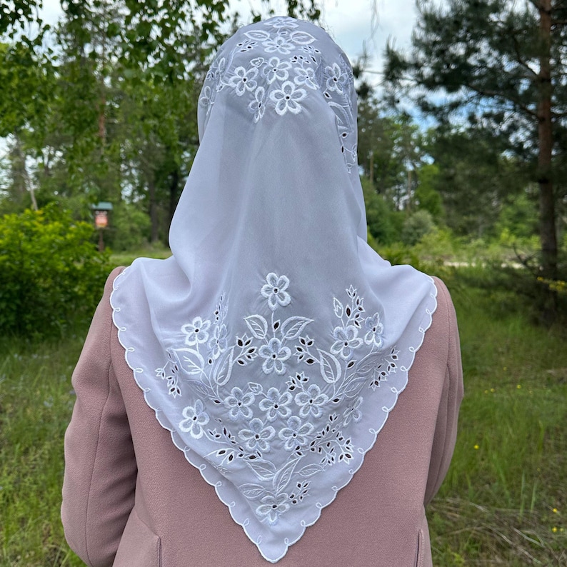 May include: A white, square head covering with intricate floral embroidery. The delicate design features detailed flowers and leaves, creating a lace-like effect. The head covering is worn over the head and shoulders, with a scalloped edge.