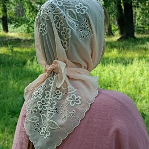 May include: A sheer, light peach-colored headscarf with intricate white floral embroidery. The scarf is tied at the nape of the neck and has a scalloped edge. The background is a blurred view of a green, grassy area.