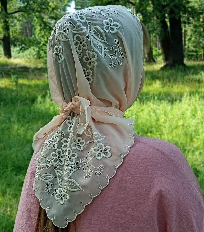 May include: A sheer, light peach-colored headscarf with intricate white floral embroidery. The scarf is tied at the nape of the neck and has a scalloped edge. The background is a blurred view of a green, grassy area.